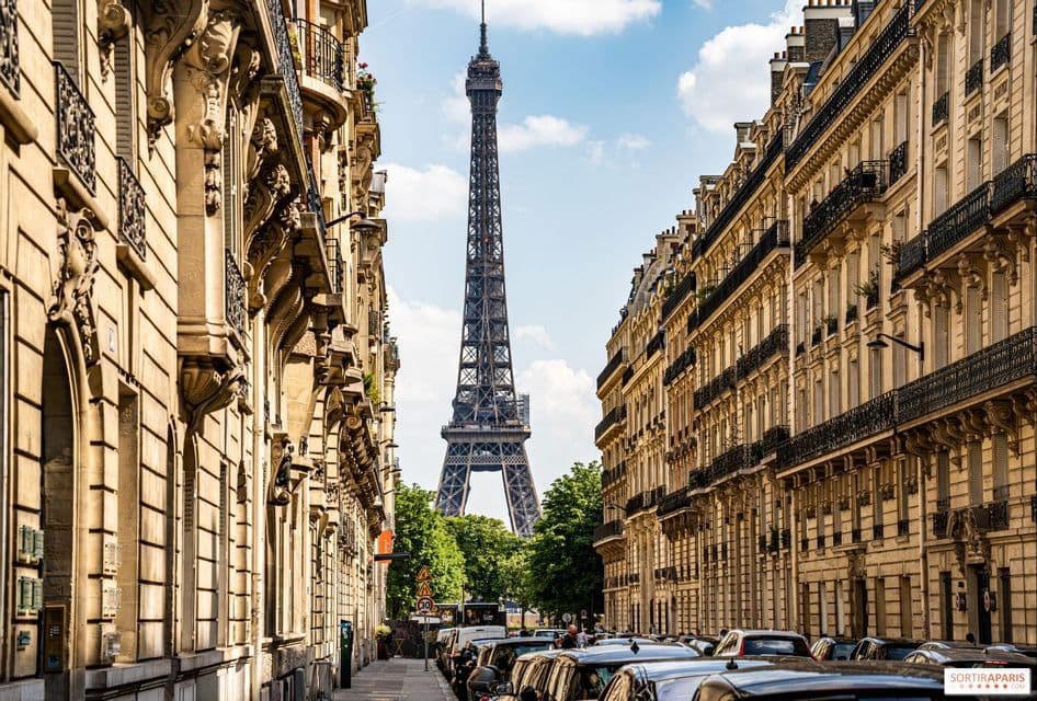 The Eiffel Tower is seen in the distance between two rows of classic apartment buildings on a street lined with cars.
