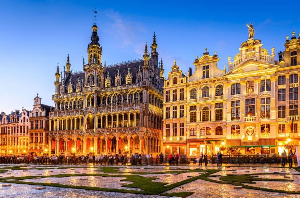A grand European square at dusk, with ornate historic buildings and a town hall brightly illuminated against a blue sky.