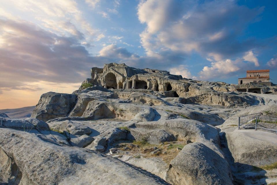 Una antigua ciudad troglodita excavada en una gran colina rocosa con un edificio de piedra en la cima, vista al atardecer.