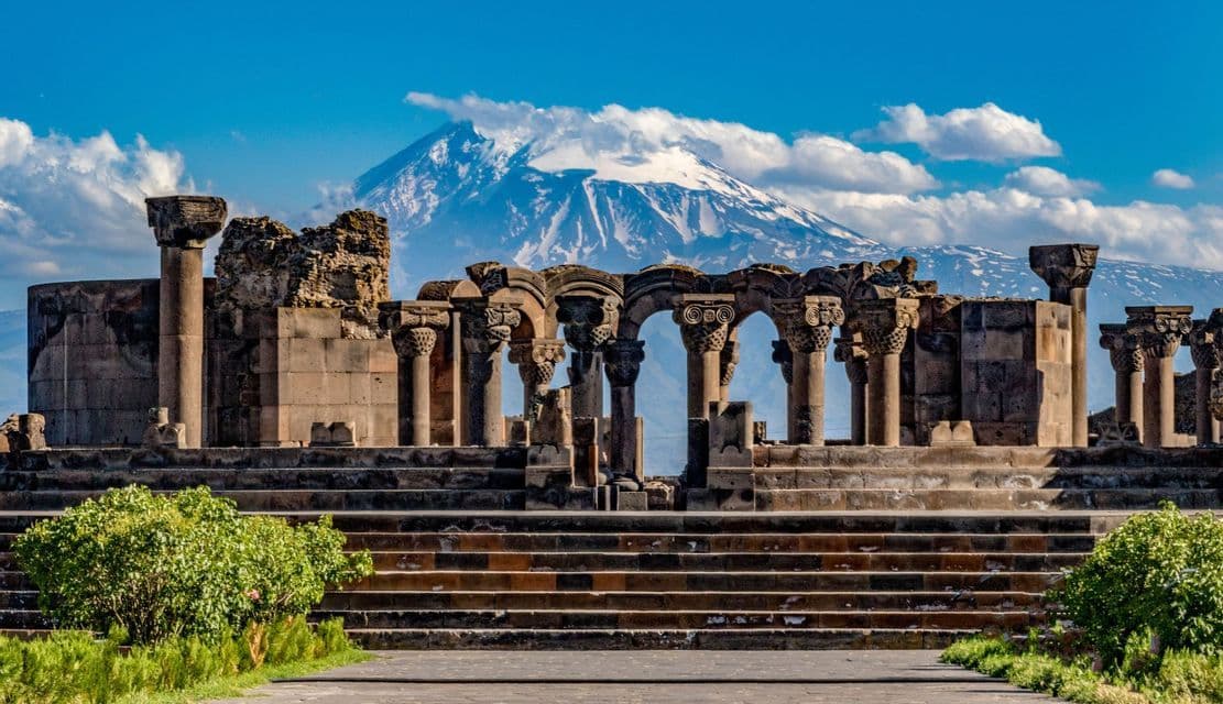 Las ruinas de un templo de piedra con columnas ornamentadas se alzan frente a una gran montaña nevada bajo un cielo azul parcialmente nublado.