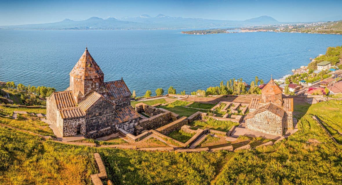 Un histórico complejo monástico de piedra en una colina verde, con vistas a un gran lago azul y montañas distantes en el horizonte bajo un cielo despejado.