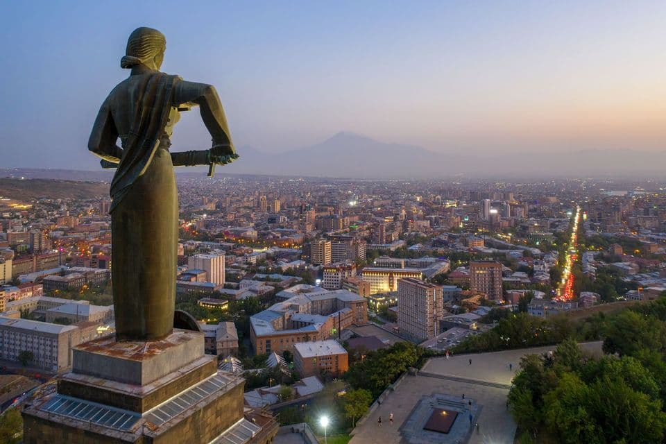 Eine große, von hinten gesehene Statue überblickt eine weitläufige, beleuchtete Stadt in der Abenddämmerung, mit einem entfernten Berg am Horizont.