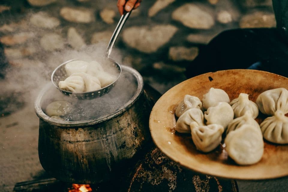 Una persona saca dumplings humeantes de una olla grande con agua hirviendo sobre el fuego, junto a un plato de madera con dumplings cocidos.