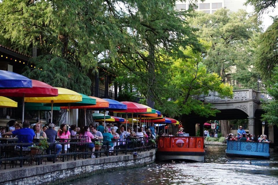 Tour boats with passengers float down a river past an outdoor restaurant with colorful umbrellas.