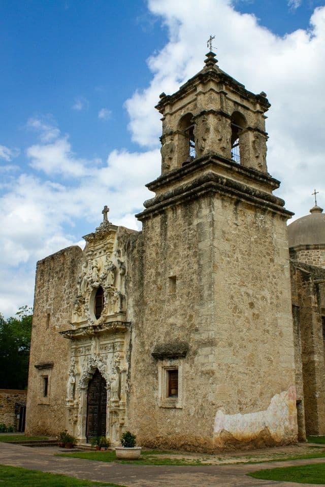 A historic stone mission with an ornate facade and a bell tower stands against a blue sky with scattered clouds.