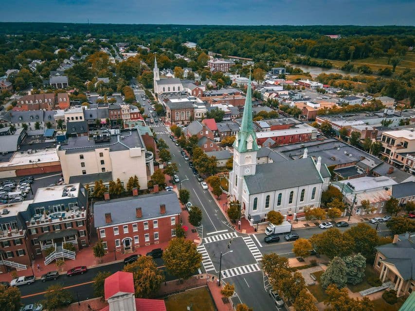 An aerial view of a historic town center with churches, brick buildings, and tree-lined streets next to a large forest and river.