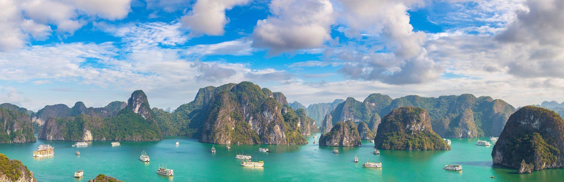 Une vue panoramique de bateaux touristiques naviguant sur des eaux turquoises parmi d'imposantes formations rocheuses couvertes de végétation sous un ciel partiellement nuageux.