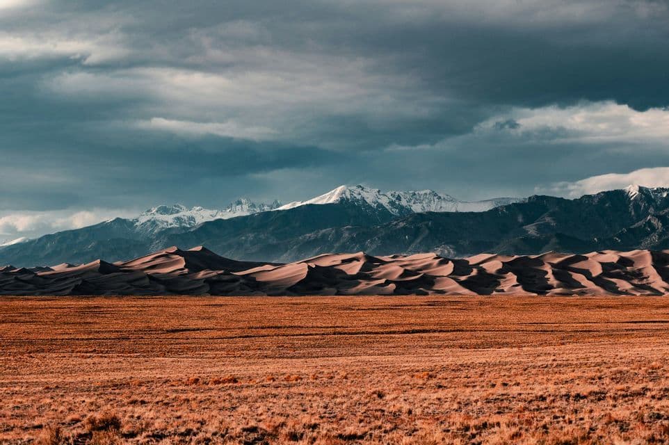 A dry grassy plain leads to rolling sand dunes with snow-capped mountains in the background under a cloudy sky.