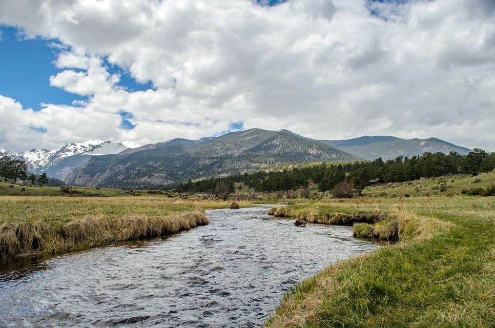 A river flows through a green meadow towards distant snow-capped mountains under a cloudy sky.