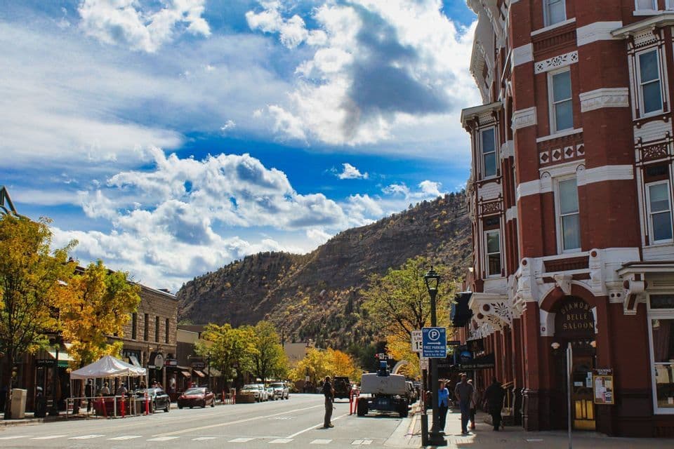 A wide street in a historic town lined with brick buildings and trees with yellow leaves, with a mountain in the background under a blue, cloudy sky.