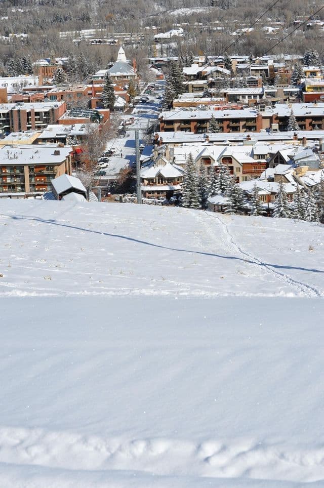 A high-angle view of a mountain town with snow-covered buildings and trees, seen from a snowy slope in the foreground.