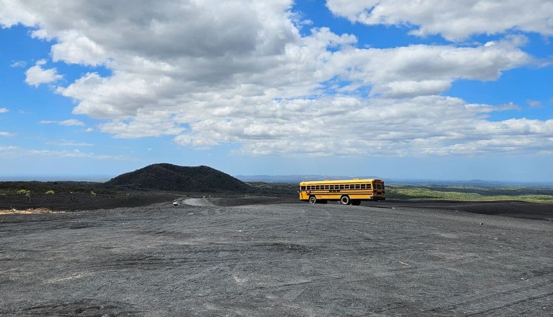 Ein gelber Schulbus steht in einer weiten, dunklen Vulkanlandschaft unter blauem Himmel mit vereinzelten weißen Wolken.