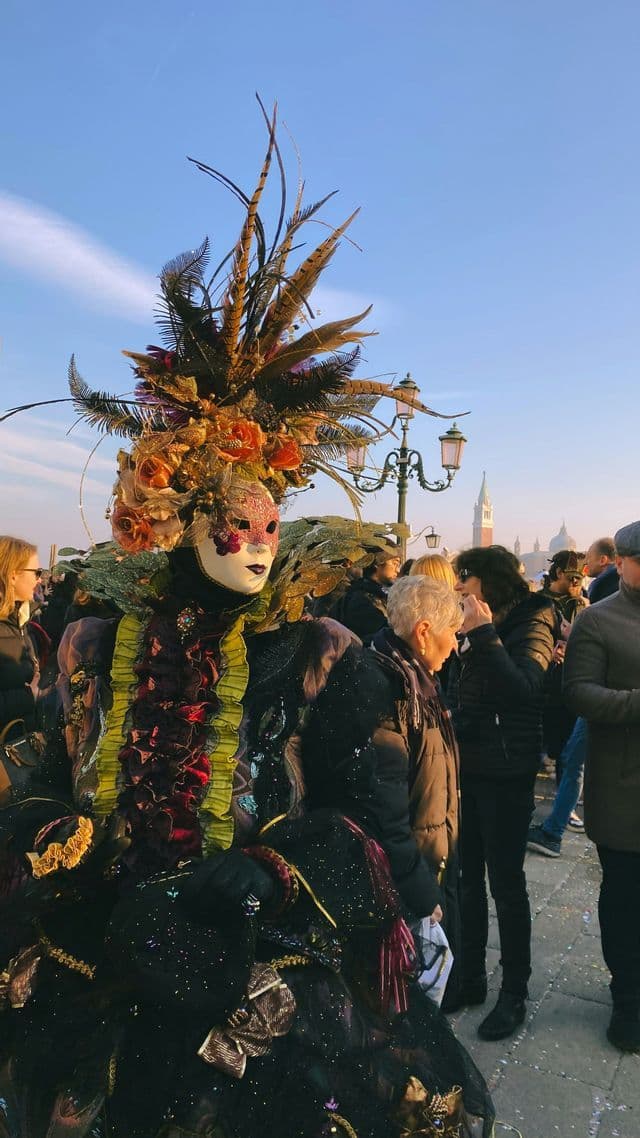 Una persona in un elaborato costume di carnevale con una maschera e un grande copricapo piumato si trova tra la folla sotto un cielo azzurro e limpido.