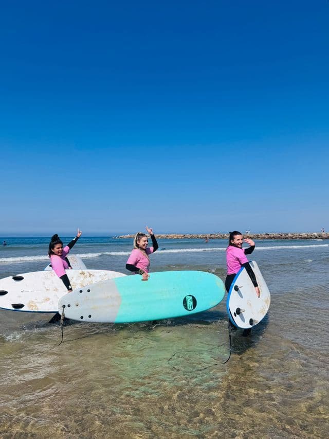 Tre donne in viaggio di gruppo WeRoad, sorridenti e in posa con le loro tavole da surf, mentre sono in piedi nell'oceano poco profondo.
