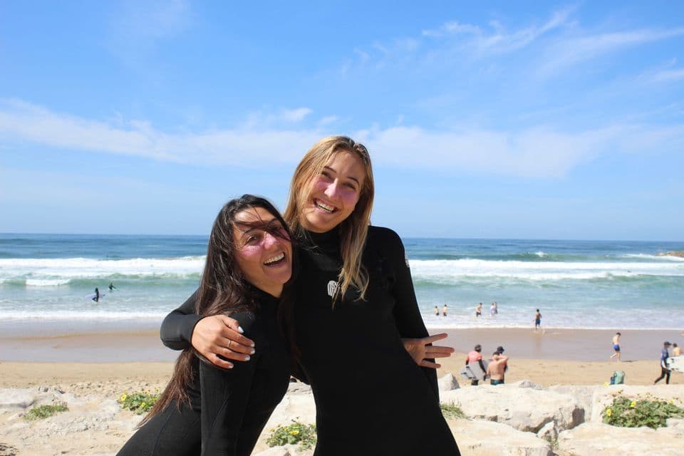 Due donne in mute nere sorridono e si abbracciano su una spiaggia rocciosa, con l'oceano e le onde sullo sfondo.