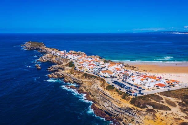 Una vista aerea di un villaggio costiero con edifici bianchi su una penisola rocciosa, delimitato dall'oceano blu profondo e da una spiaggia sabbiosa.