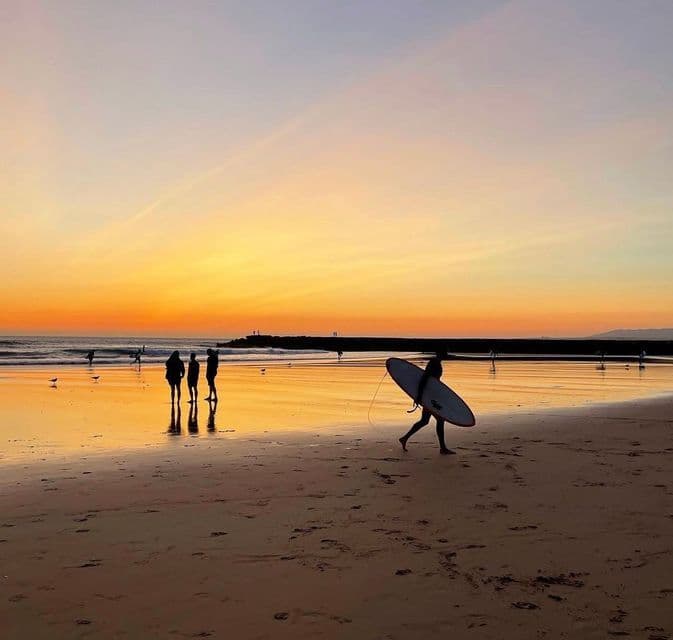 Un surfista in controluce porta una tavola da surf lungo una spiaggia bagnata al tramonto, con altre persone sullo sfondo.