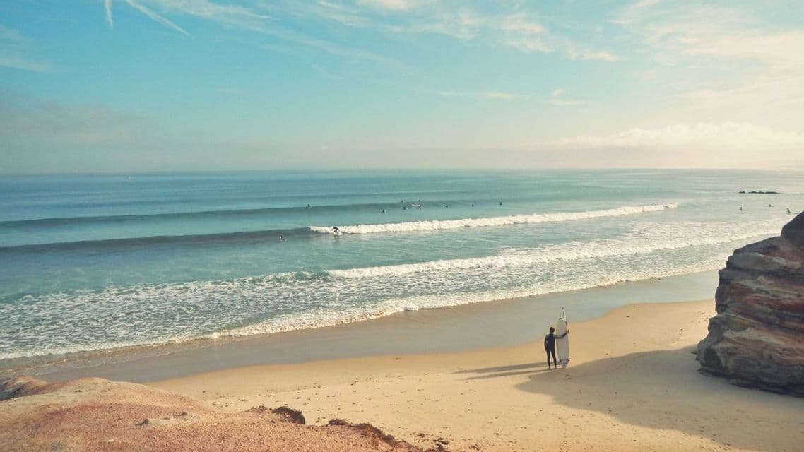 Un surfista con una tavola da surf è su una spiaggia sabbiosa, osservando altri surfisti sulle onde dell'oceano.