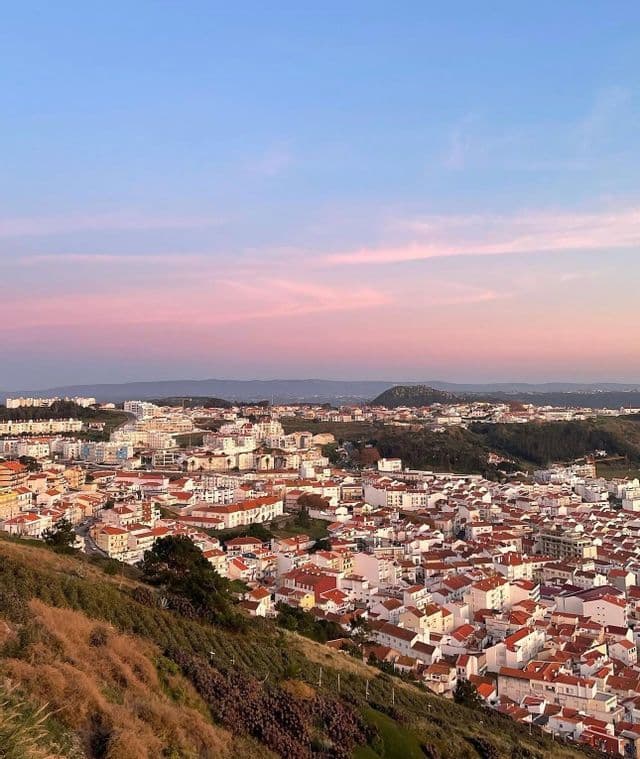 Un fitto paesaggio urbano con edifici bianchi e tetti rossi, visto da una collina sotto un cielo rosa e azzurro al tramonto.