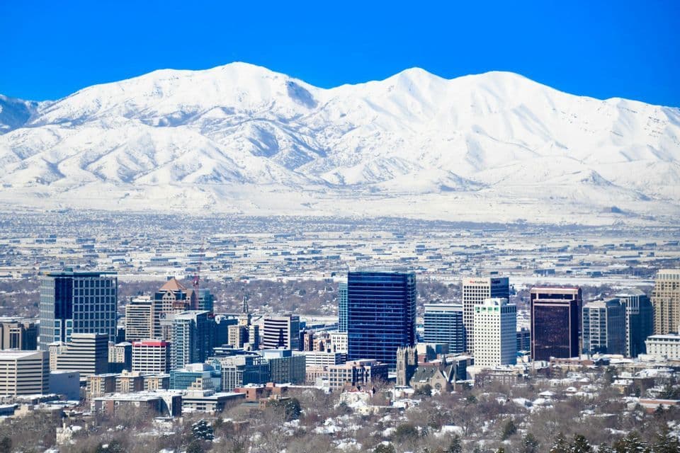A dense city skyline stands before a vast, snow-covered mountain range under a clear blue sky.