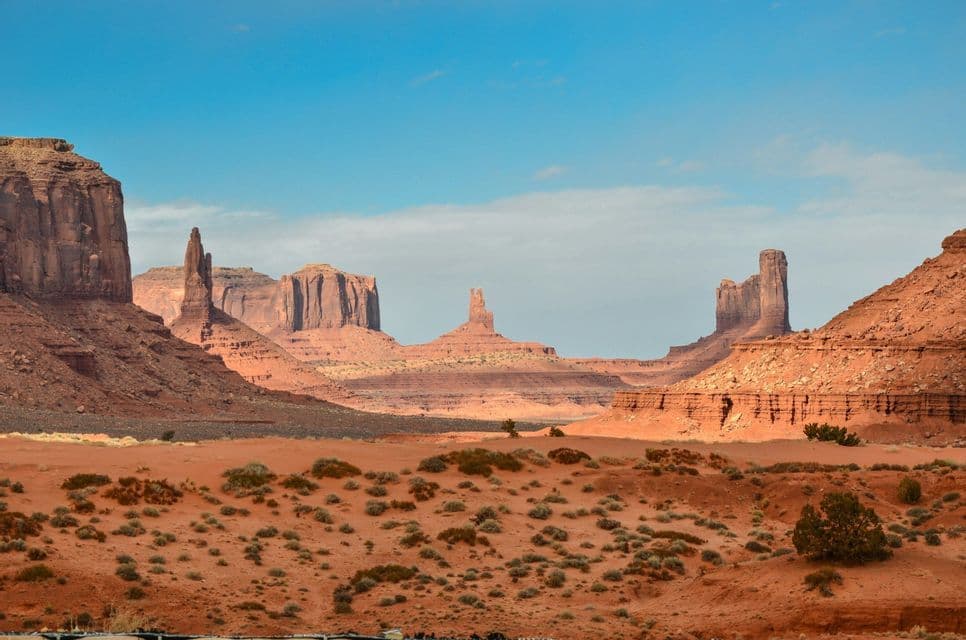 A wide desert valley with red sandstone buttes and mesas rising in the distance under a partly cloudy blue sky.