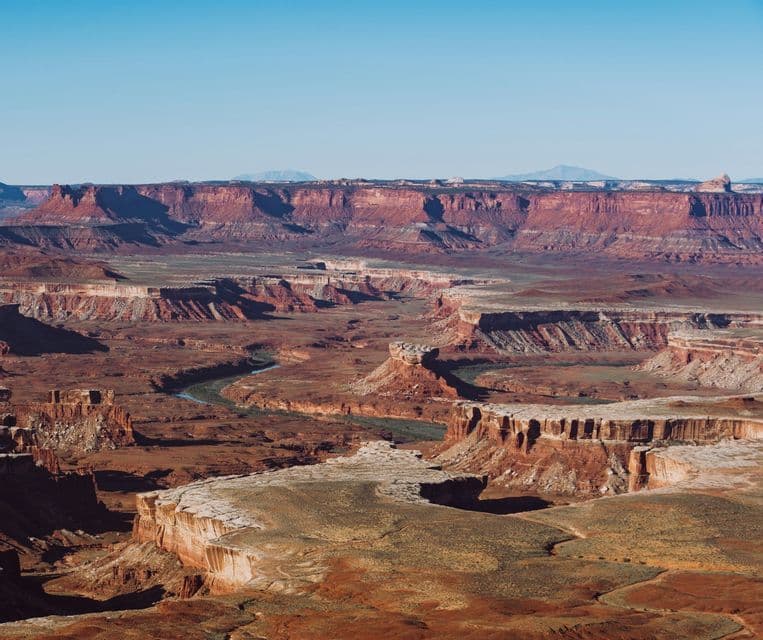 A wide, sunny view of a river snaking through a vast landscape of red rock canyons and mesas under a clear sky.