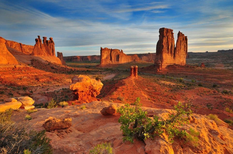 Red rock formations rise from a desert landscape, illuminated by warm sunrise light under a blue, partly cloudy sky.