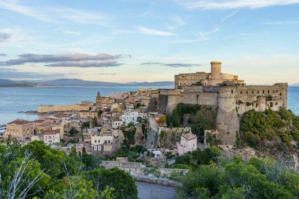 Una grande fortezza di pietra si erge su una scogliera sopra una città costiera, con il mare e le montagne lontane sullo sfondo.