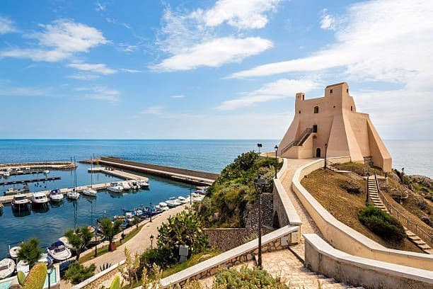 Una fortezza su una scogliera costiera domina un porticciolo pieno di barche sotto un cielo blu con nuvole bianche.