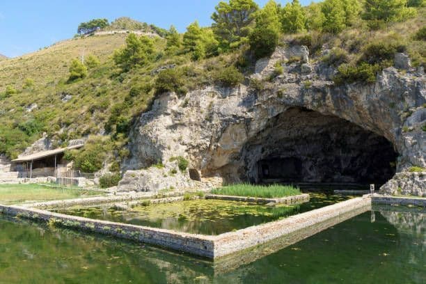 Una grande grotta alla base di un'altura rocciosa e verdeggiante, con una piscina d'acqua rettangolare in pietra in primo piano.