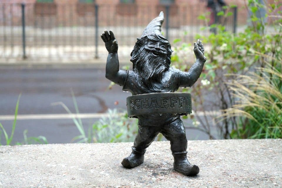 Una piccola statua di metallo scuro raffigurante uno gnomo sorridente con le braccia alzate si trova su un bordo di cemento bagnato, con una strada sfocata sullo sfondo.