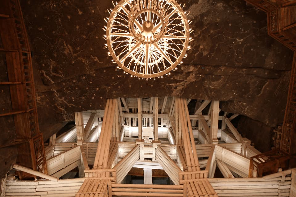 Una vista dal basso di un grande lampadario illuminato, sospeso da un soffitto roccioso e scuro sopra un'intricata struttura in legno, in una caverna.