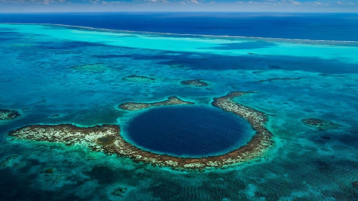 An aerial view of a large, circular, dark blue marine sinkhole surrounded by a coral atoll in a turquoise ocean.