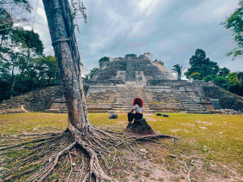 A person with red hair sits on a rock in a grassy clearing, looking at a large, tiered stone pyramid surrounded by jungle trees.