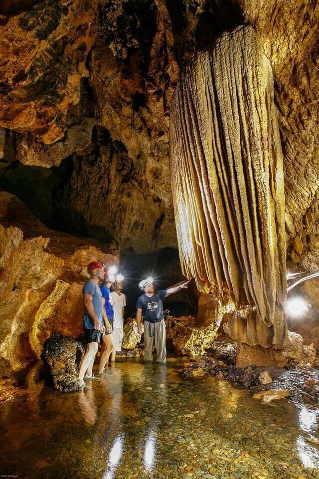 A guide points at a large stalactite for a WeRoad group trip exploring a cave with headlamps while standing in water.