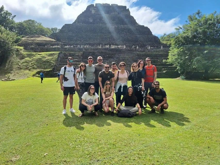 A WeRoad group trip poses for a photo on a grassy lawn in front of a large stone step pyramid under a partly cloudy sky.