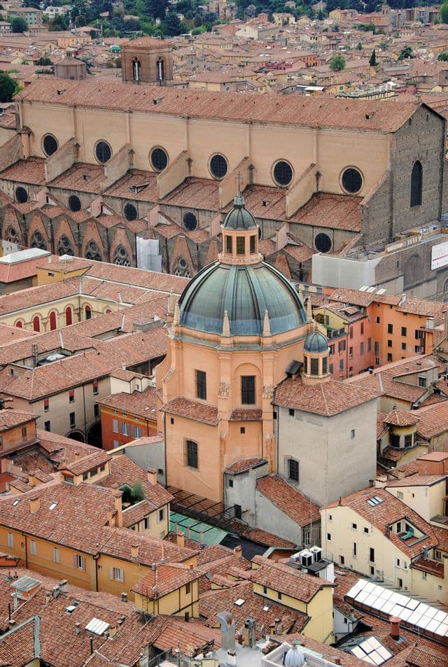 An aerial view of a historic city with red-tiled roofs, featuring a prominent green dome and a large basilica.