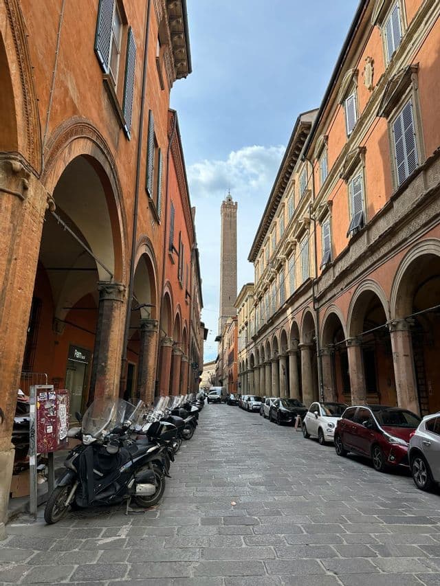 A narrow cobblestone street lined with porticoes, with parked scooters and a tall tower visible in the distance.