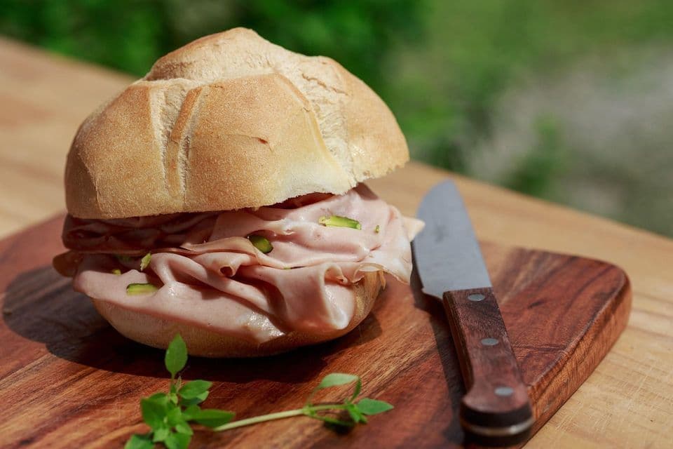 A mortadella sandwich in a bread roll sits on a wooden cutting board next to a small knife and a sprig of herbs.