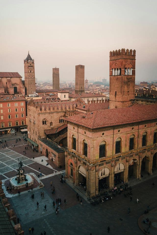 An aerial view of a historic European plaza with brick towers and terracotta-roofed buildings as people walk below during a hazy sunset.