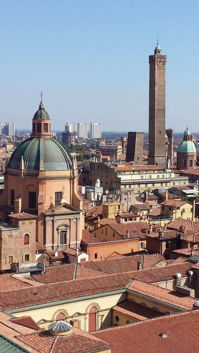 An aerial view of a historic European city, featuring red terracotta roofs, a green-domed church, and a tall brick tower.