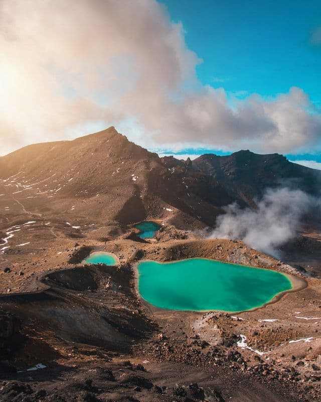 An aerial view of three bright turquoise crater lakes set in a barren volcanic mountain landscape with steam rising nearby.
