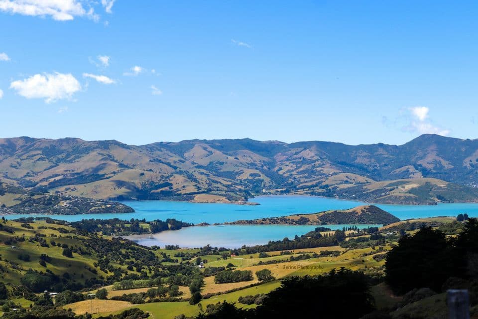 A high-angle view of a turquoise bay surrounded by rolling green and brown hills under a partly cloudy blue sky.