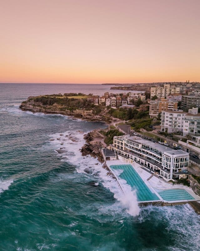 An aerial view of large ocean waves crashing against a seaside swimming pool on a rocky coastline during sunset.