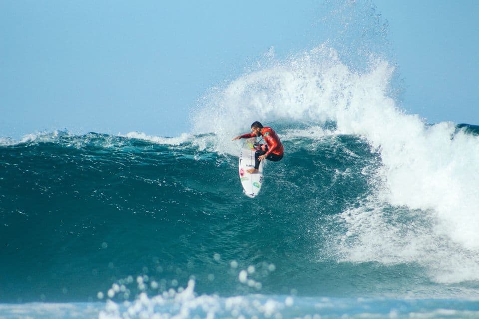 A surfer in a red wetsuit rides the crest of a large, breaking ocean wave under a clear blue sky.