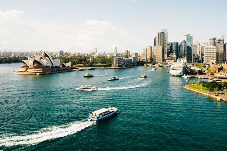 An aerial view of ferries cruising on turquoise water in front of the Sydney Opera House and a modern city skyline.