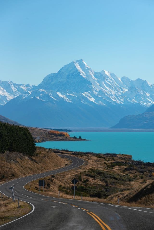 A winding road curves through a hilly landscape beside a turquoise lake, with snow-capped mountains in the distance.