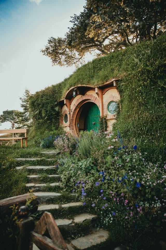 A hobbit-style home with a round green door built into a grassy hill, with stone steps leading up through a garden of flowers.