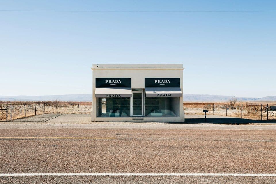 The Prada Marfa art installation, a standalone storefront, sits by an empty road in the desert under a clear blue sky.