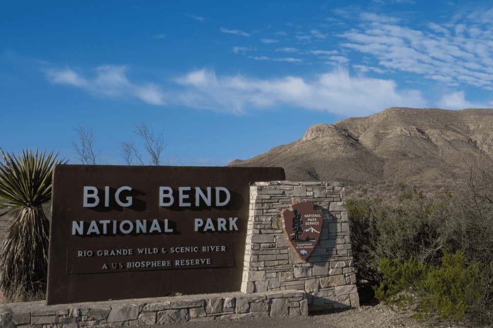 The entrance sign for Big Bend National Park stands in a desert landscape with a mountain in the background under a blue sky.